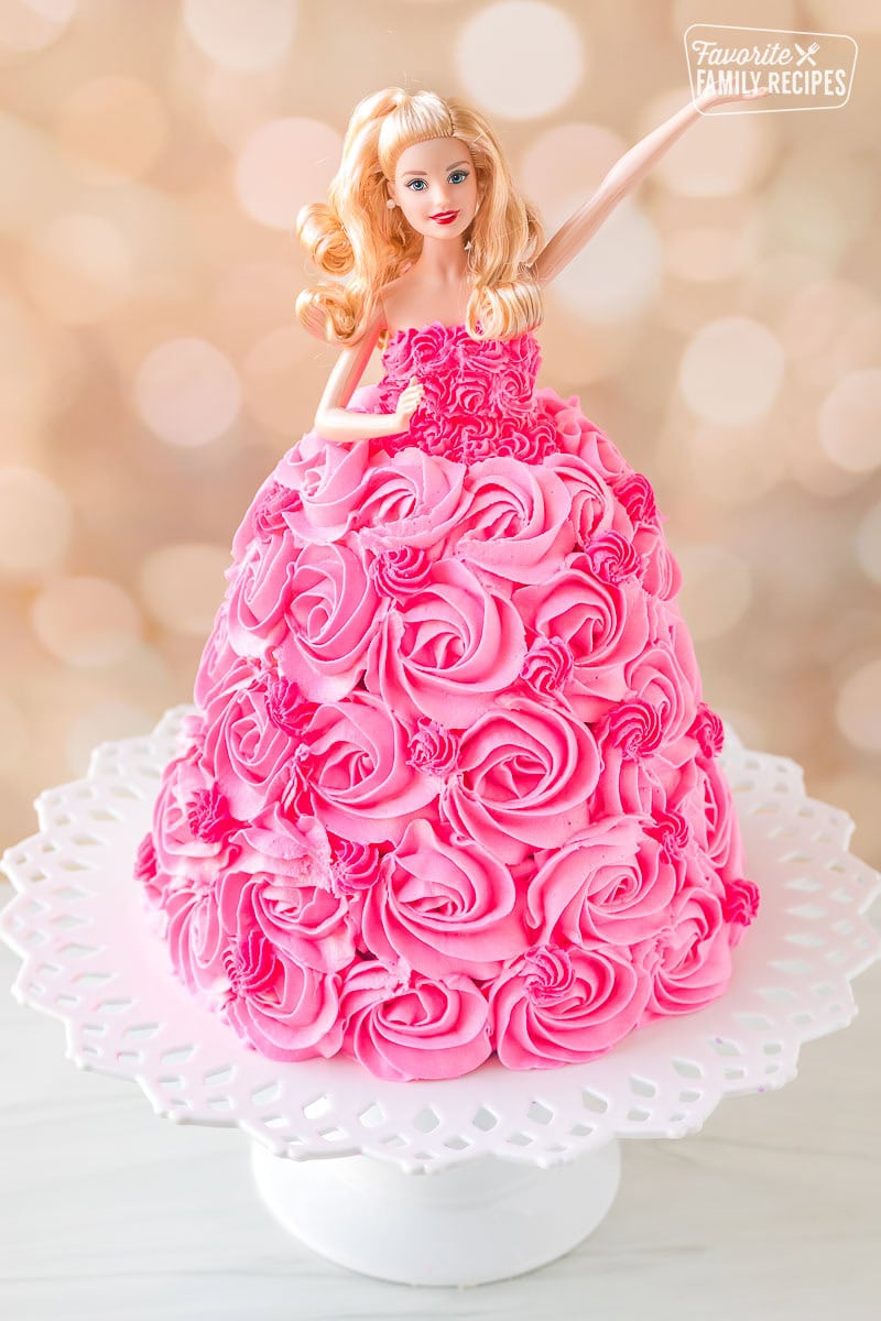 Barbie cake on a cake stand with her arm lifted up.