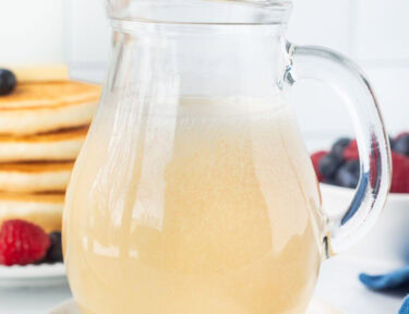 A glass pitcher filled with homemade buttermilk syrup with a stack of pancakes in the background.