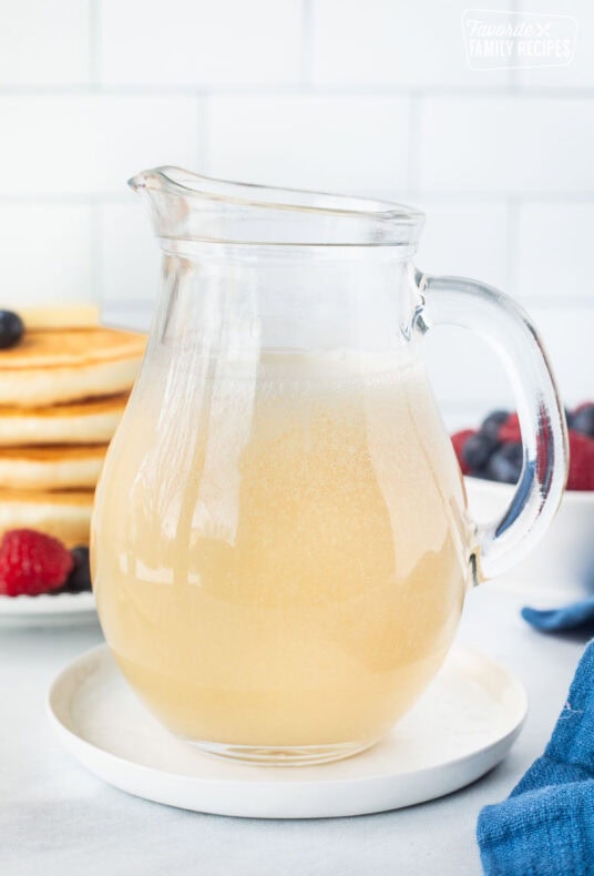 A glass pitcher filled with homemade buttermilk syrup with a stack of pancakes in the background.