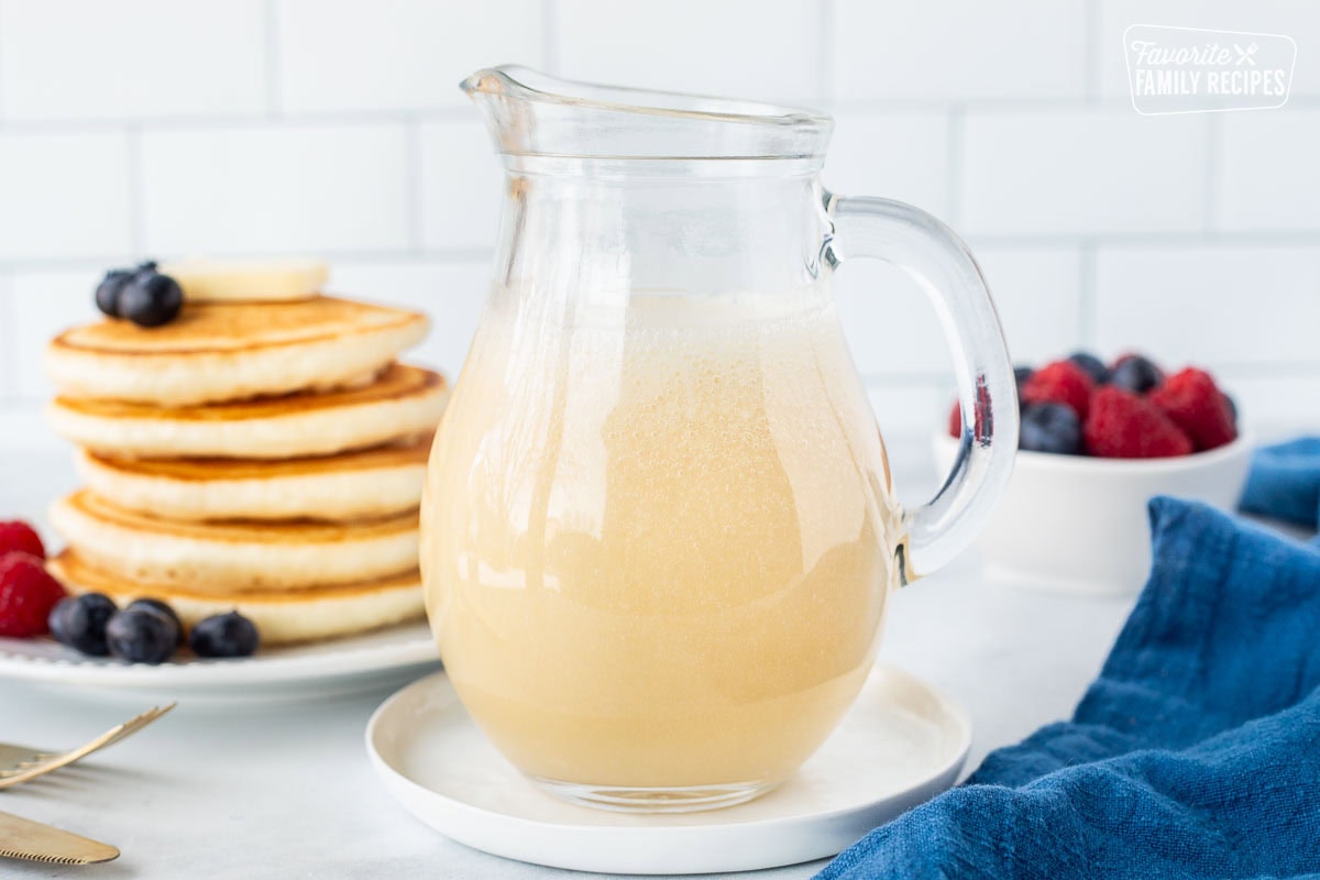 A clear glass pitcher filled with homemade buttermilk syrup sits on a white saucer. Behind it is a plate of golden brown pancakes topped with blueberries and sliced bananas. A small white bowl filled with fresh raspberries and blueberries is also visible. A blue napkin is draped on the right side. The "Favorite Family Recipes" logo is in the upper right corner.