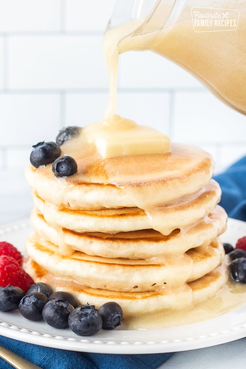 Creamy buttermilk syrups being poured onto a stack of buttermilk pancakes with fresh blueberries and raspberries on the side of the plate.