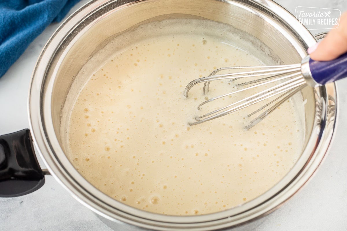 A hand holding a whisk is stirring homemade buttermilk syrup simmering in a stainless steel saucepan. 