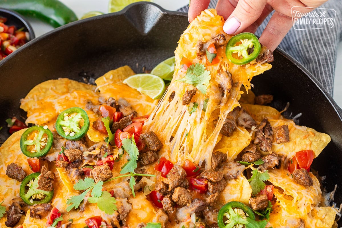 A cheesy nacho topped with savory carne asada, cilantro, and tomatoes being pulled from a skillet of nachos showing the delicious cheese pulling up from the rest of the stack. 