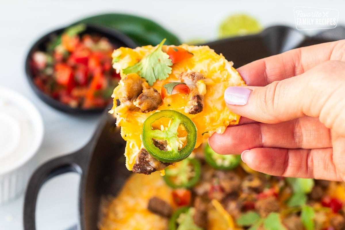 Hand holding up a Carne Asada nacho with melted cheese pieces of Carne Asada, cilantro and jalapeño showing all the delicious textures of all the savory ingredients. 