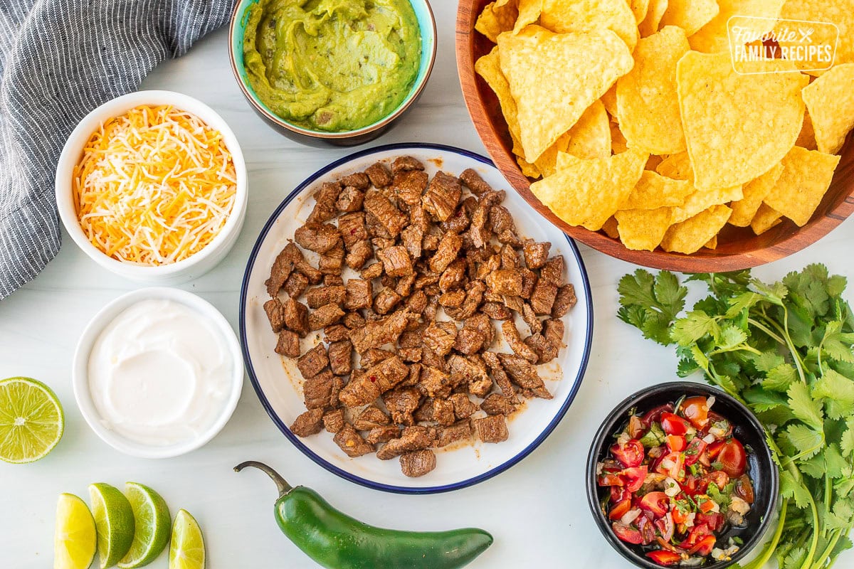 Ingredients to make Carne Asada Nachos including tortilla chips, Carne Asada, pico de gallo, cilantro, jalapeño, lime wedges, sour cream, shredded cheddar cheese and guacamole.