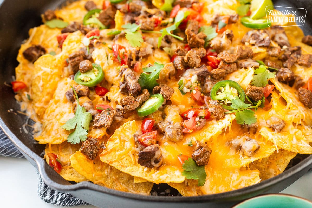 Carne Asada nachos with toppings including savory, seasoned carne asada bites, sliced jalapeños, pico de gallo, cilantro, and lime wedges.