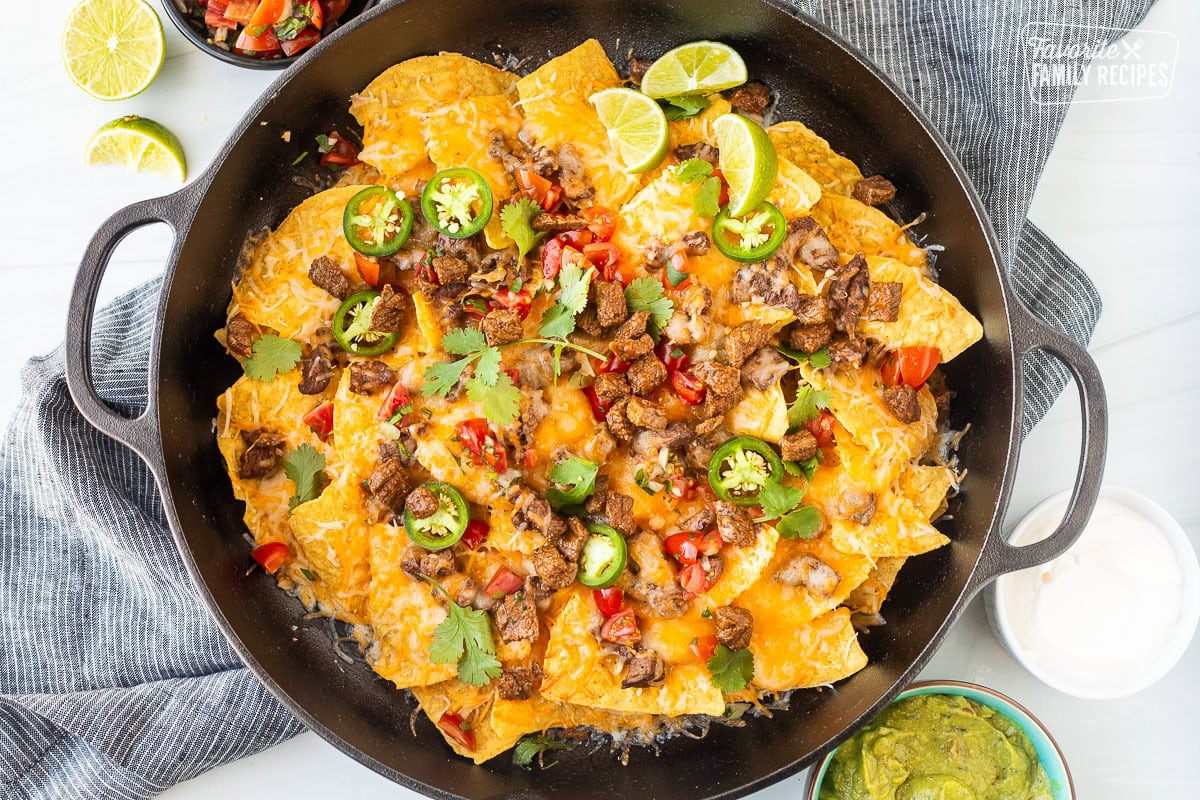 Carne Asada nachos with toppings including seasoned, diced carne asada, sliced jalapeños, pico de gallo, cilantro, and lime wedges.