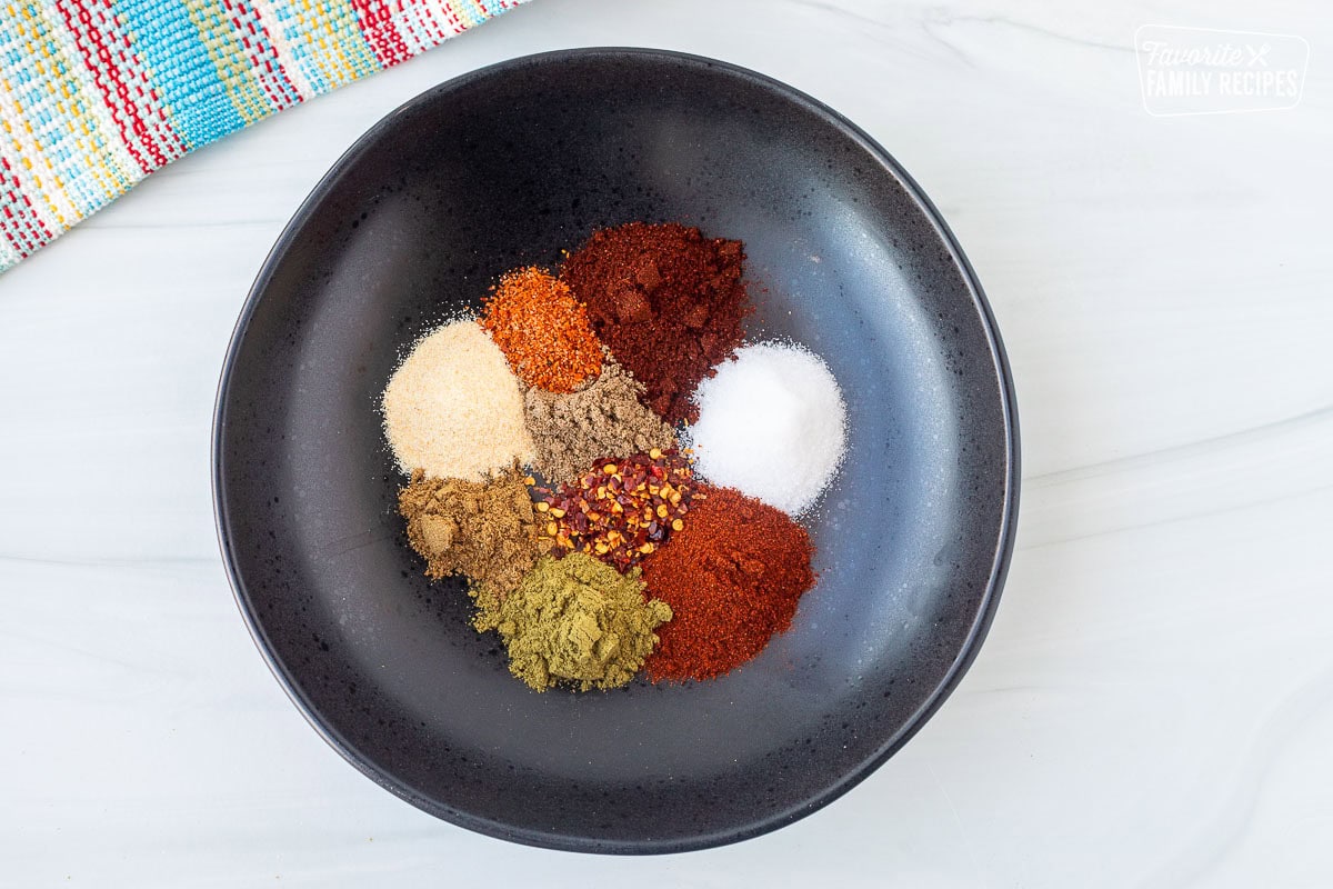 Glass bowl with measured garlic powder, onion powder, cumin, oregano, salt, coriander, red pepper flakes, chili powder, chipotle chili Pepper powder and Tajin.