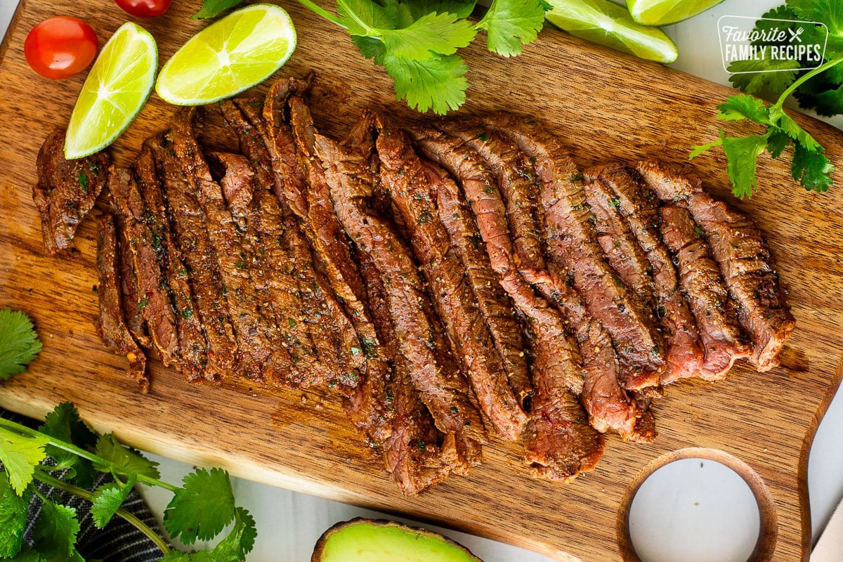 Grilled, juicy, Carne Asada sliced on a wooden cutting board seasoned with carne asada seasoning surrounded by fresh limes, cilantro, and tomatoes.