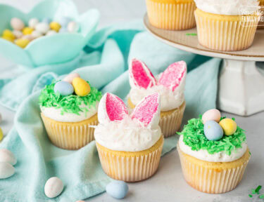 Decorated Easter cupcakes with two designs. One design has bunny ears made with marshmallows and the other has a coconut nest of chocolate Cadberry eggs.