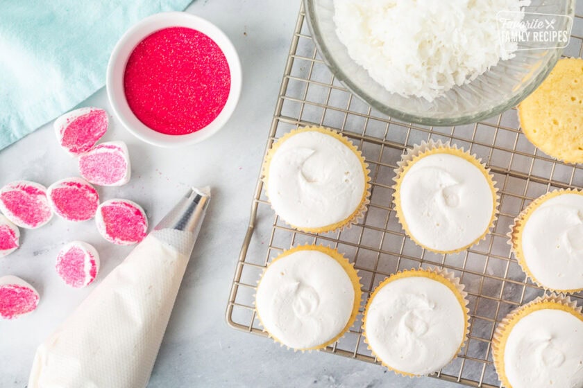 Frosted cupcakes with a decorating tip next to a bowl of shredded coconut and pink sprinkles and marshmallows shaped like bunny ears.