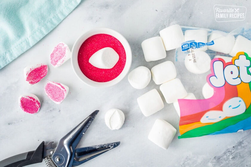 Large marshmallows cut in half with scissors and dipped into a bowl of pink sprinkles.