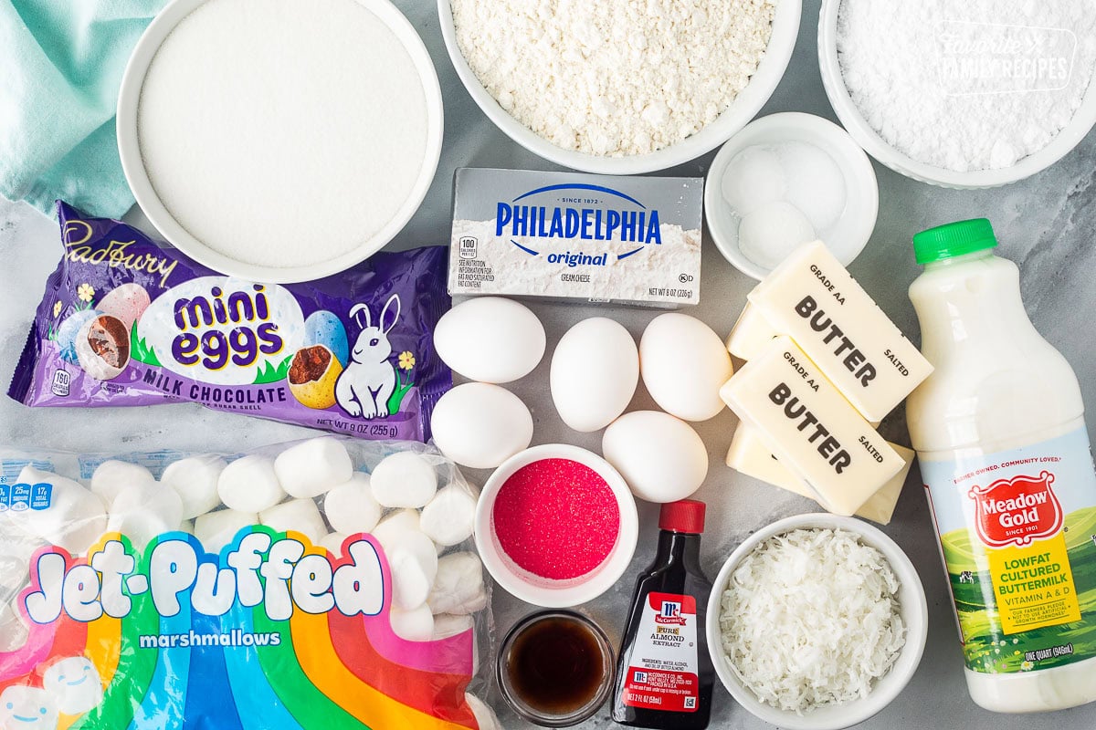 Ingredients to make Easter cupcakes including buttermilk, salt, baking powder, baking soda, powdered sugar, coconut, butter, eggs, almond extract, vanilla, sprinkles, cream cheese, flour, sugar, mini Cadberry eggs and large marshmallows.