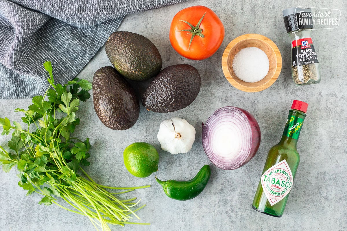 Ingredients to make guacamole including avocado, tomato, salt, pepper, red onion, jalapeño, garlic, lime, cilantro and green Tabasco sauce.