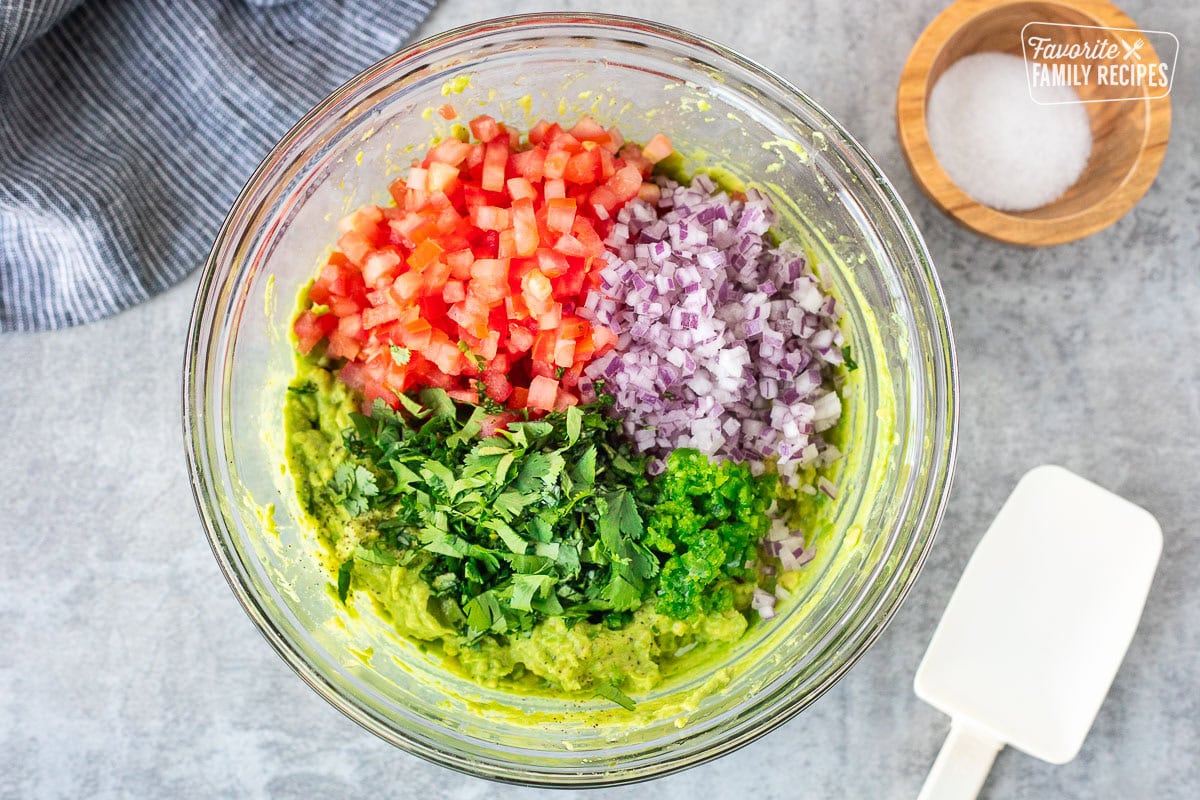 Glass mixing bowl with mash avocados and diced up tomatoes, red onions, cilantro and jalapeño on top.