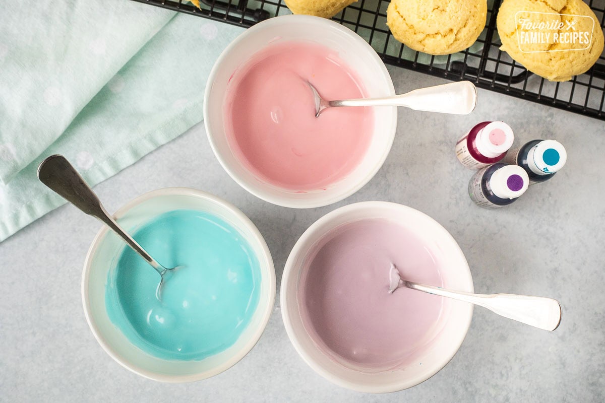 Three bowls of colored icing in pink, purple and blue.