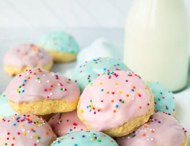 Italian Easter cookies frosted with purple, pink and blue icing and sprinkles stacked together on a plate. Glass of milk in the background.