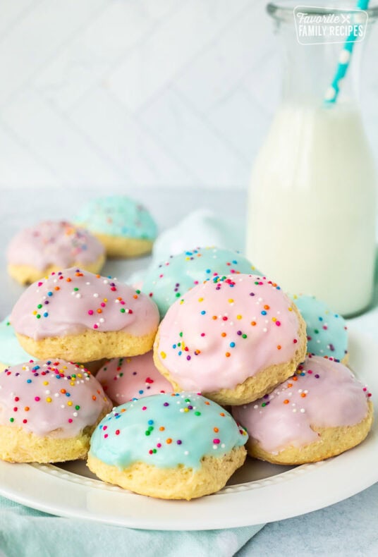 Italian Easter cookies frosted with purple, pink and blue icing and sprinkles stacked together on a plate. Glass of milk in the background.