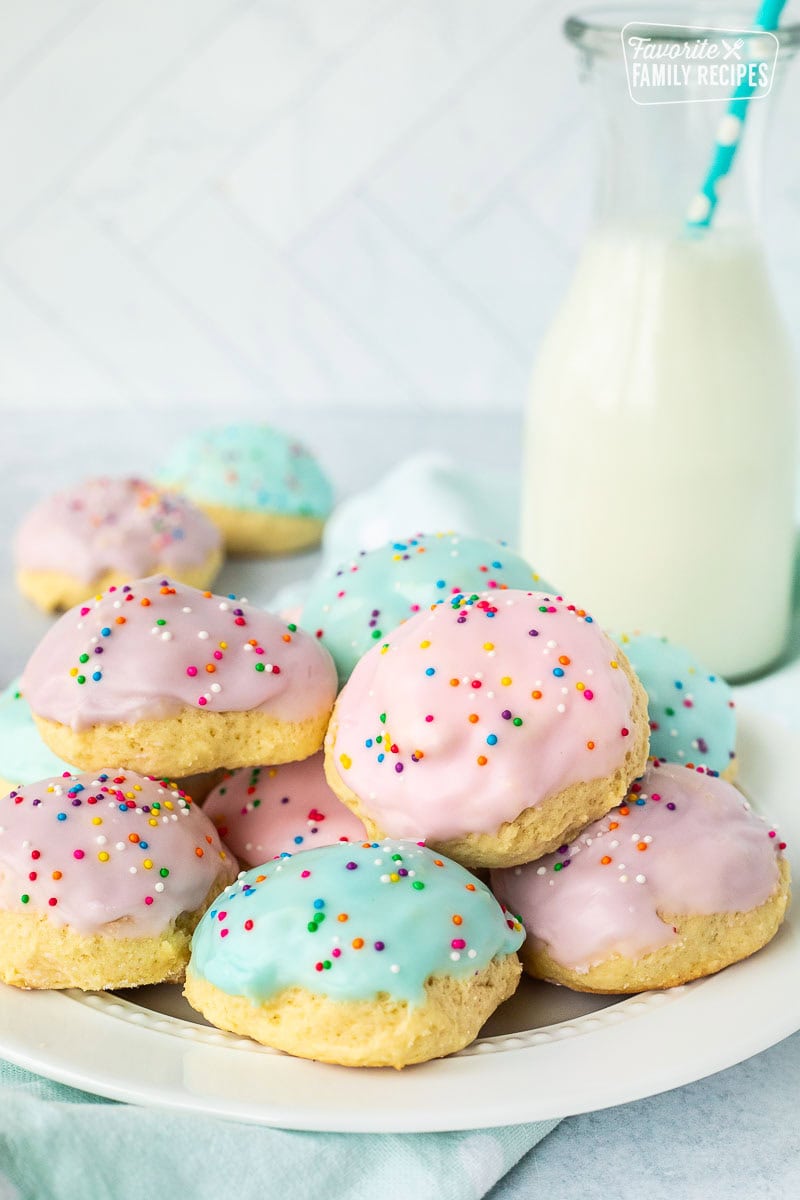 Italian Easter cookies frosted with purple, pink and blue icing and sprinkles stacked together on a plate. Glass of milk in the background.