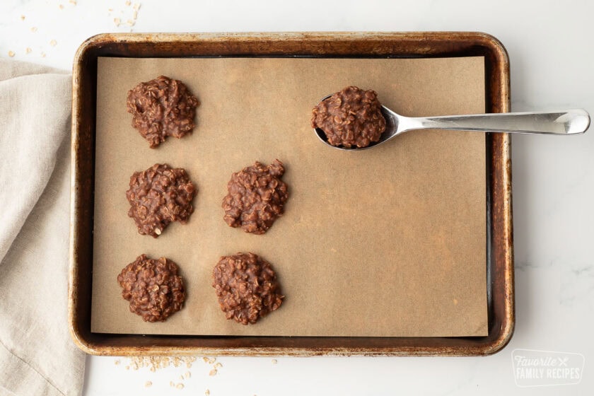 A baking sheet with scoops of chocolate no bake cookies with a spoonful being placed on the pan