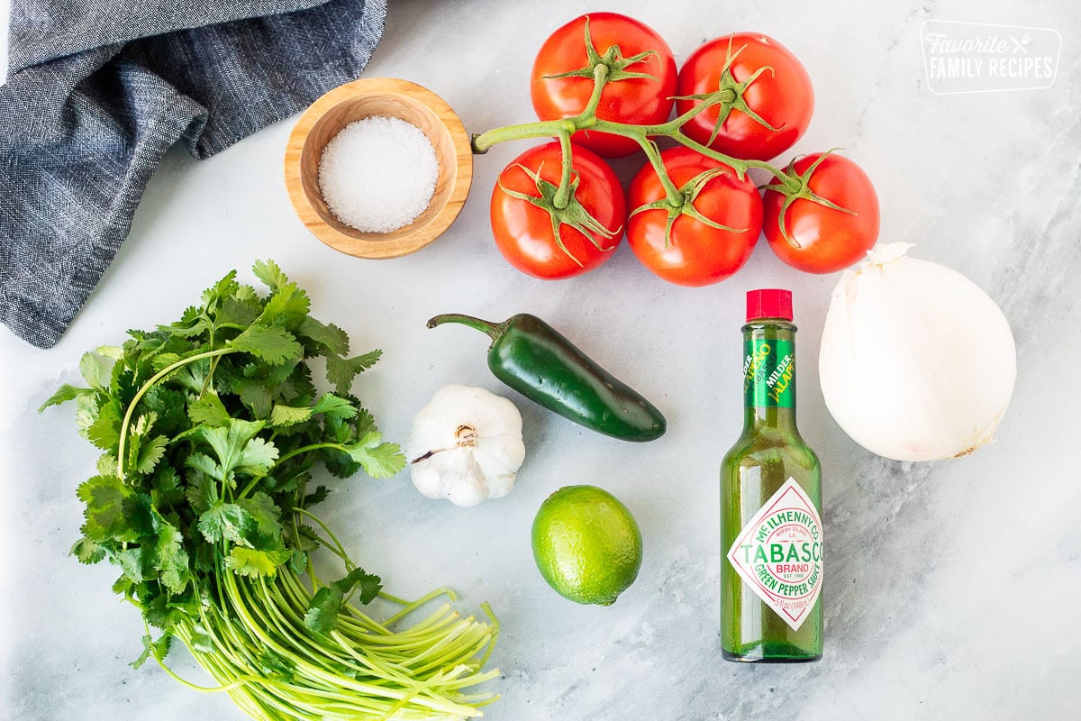 Ingredients to make Pico de Gallo including tomatoes on the vine, salt, white onion, green Tabasco sauce, lime, jalapeño, garlic and fresh cilantro.