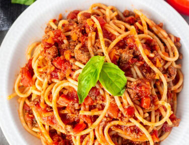 Classic spaghetti with rich Italian meat sauce garnished with fresh basil in a bowl with tomatoes and a wedge of parmesan cheese on the side.