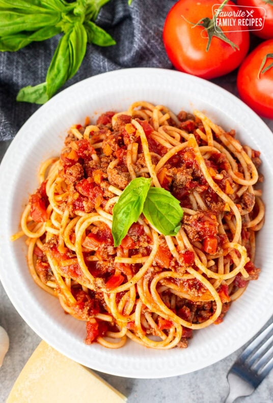 Classic spaghetti with rich Italian meat sauce garnished with fresh basil in a bowl with tomatoes and a wedge of parmesan cheese on the side.