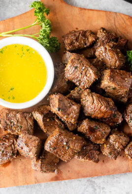 Air fryer steak bites on a wooden board with a small bowl of melted parsley butter on the side.