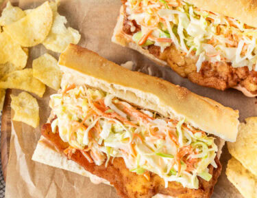 Two Cajun Fried Chicken Po'Boys on a cutting board with chips.