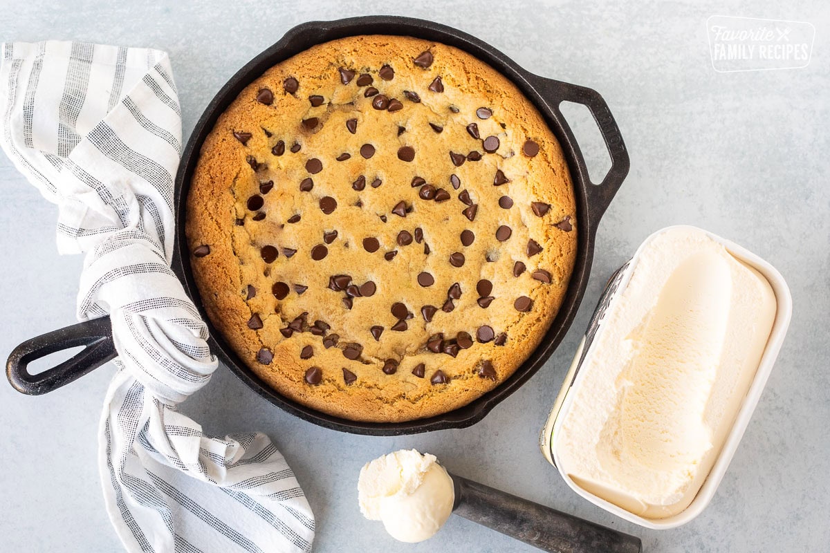 Overhead view of a chocolate chip Pizookie in a black cast iron skillet, with a scoop of vanilla ice cream on a metal scoop and a container of vanilla ice cream next to it. 