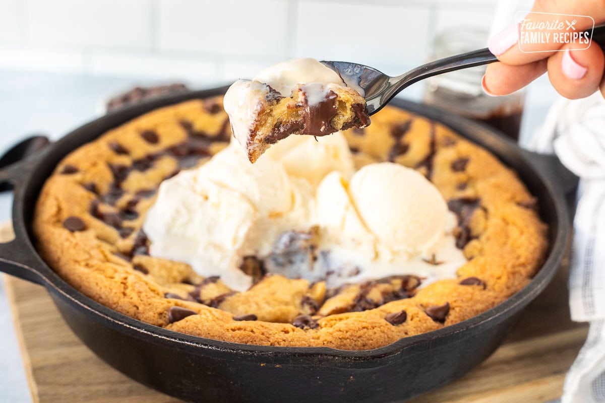 Close-up of a hand holding a spoonful of Pizookie with melting vanilla ice cream and chocolate, lifted from a cast iron skillet. The Pizookie has a golden brown crust and visible chocolate chips.