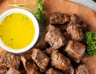 Air fryer steak bites on a wooden board with a small bowl of melted parsley butter on the side.