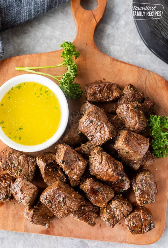 Air fryer steak bites on a wooden board with a small bowl of melted parsley butter on the side.